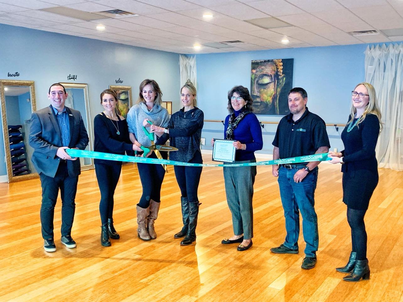 Group of people participating in a ribbon-cutting ceremony indoors with a large pair of scissors, wooden floor, and artwork on the wall.