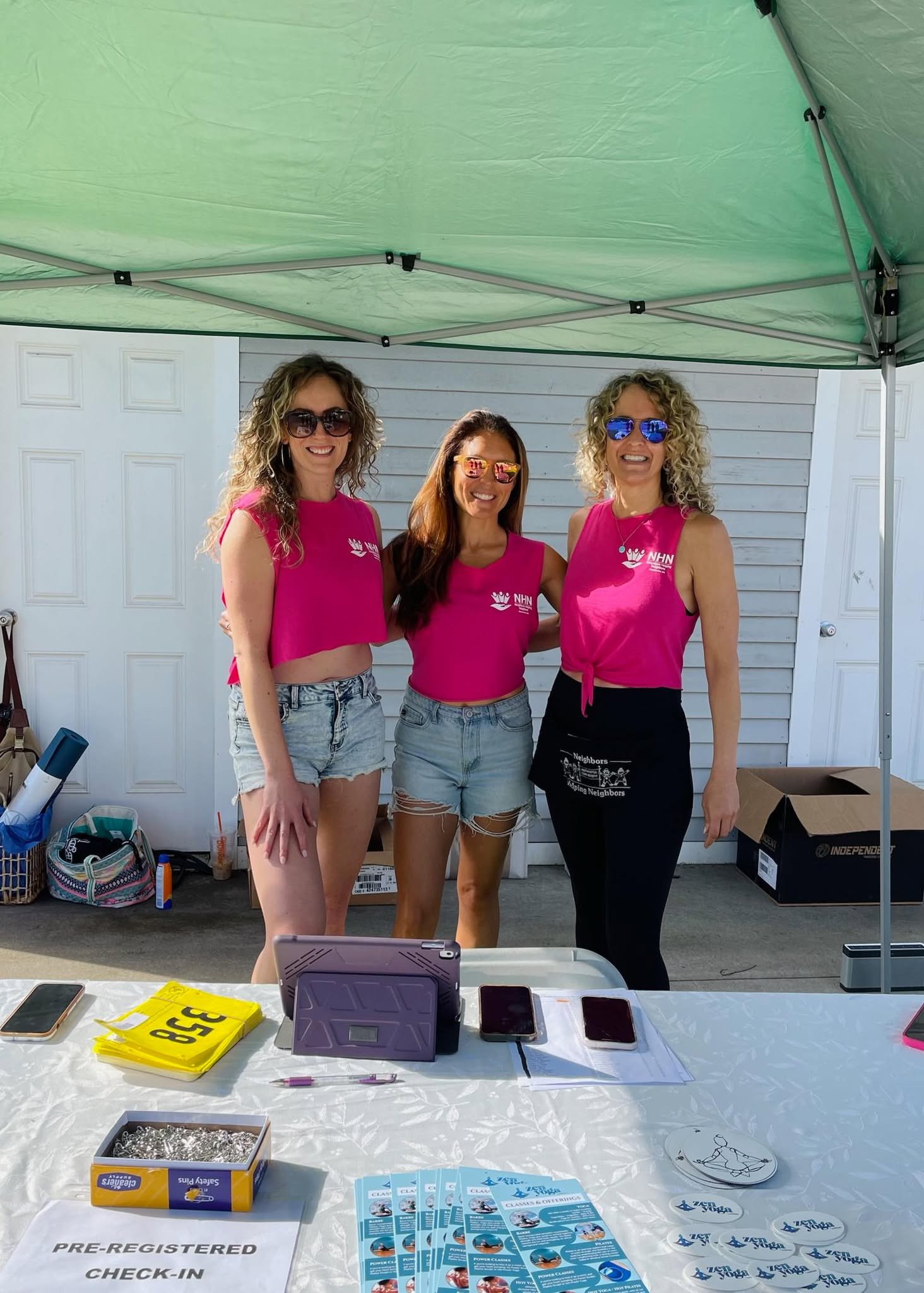 Three women in pink tank tops standing under a green canopy tent behind a check-in table with registration materials and promotional items.