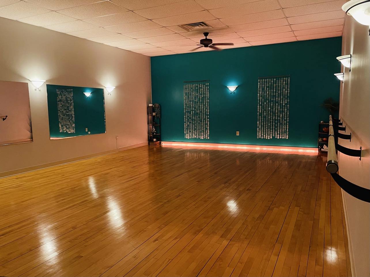 Empty dance studio with hardwood floor, teal accent wall, mirrors, ballet barres, ceiling fan, and soft overhead lighting.