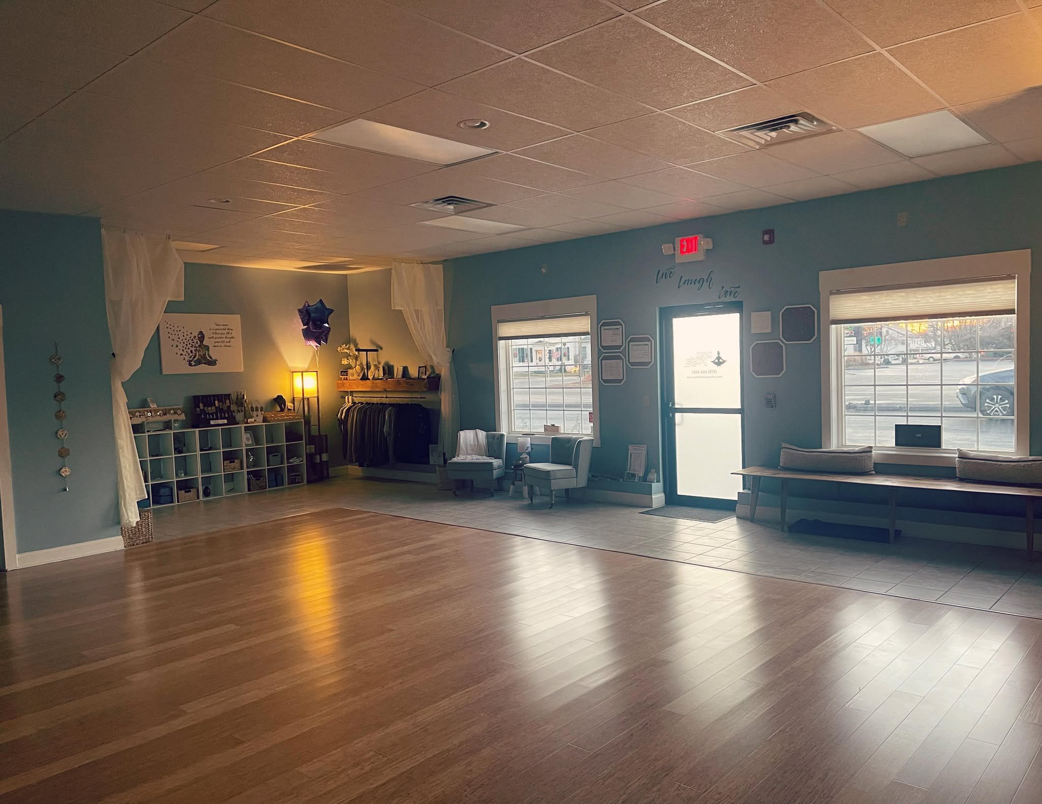 Empty yoga studio with wooden floors, light blue walls, and calm atmosphere. Shelving with yoga mats, clothing rack, and cozy seating area by windows. Wall art and decorations present.