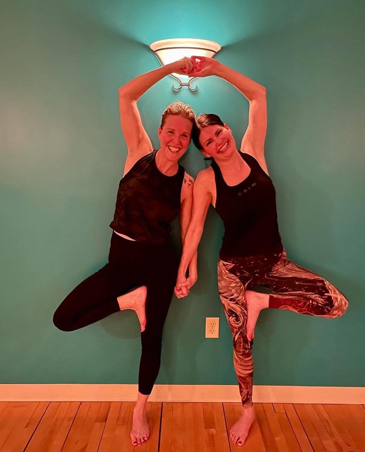 Two women performing a yoga pose, standing on one leg with the other leg bent and foot on the inner thigh, arms raised forming a heart shape. They are smiling, wearing workout attire, against a teal wall with a light fixture above.
