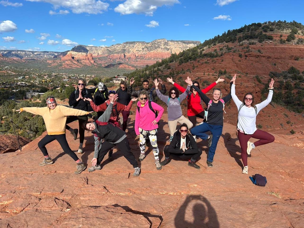A group of people posing energetically on a red rock formation with scenic desert mountains in the background. The sky is clear with a few clouds.