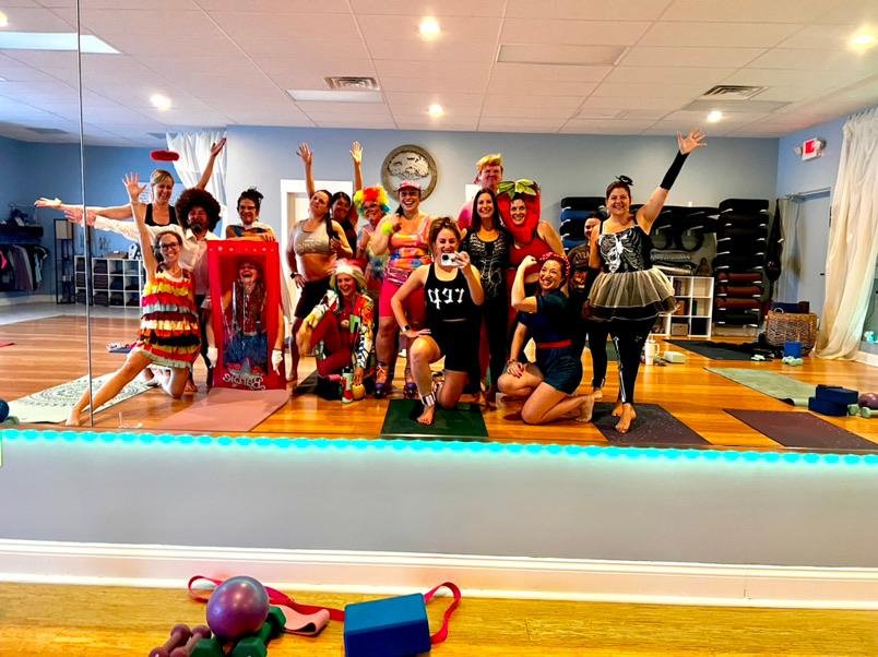 Group of people in costumes posing in a yoga studio, with colorful outfits and props, mirrored reflection visible.