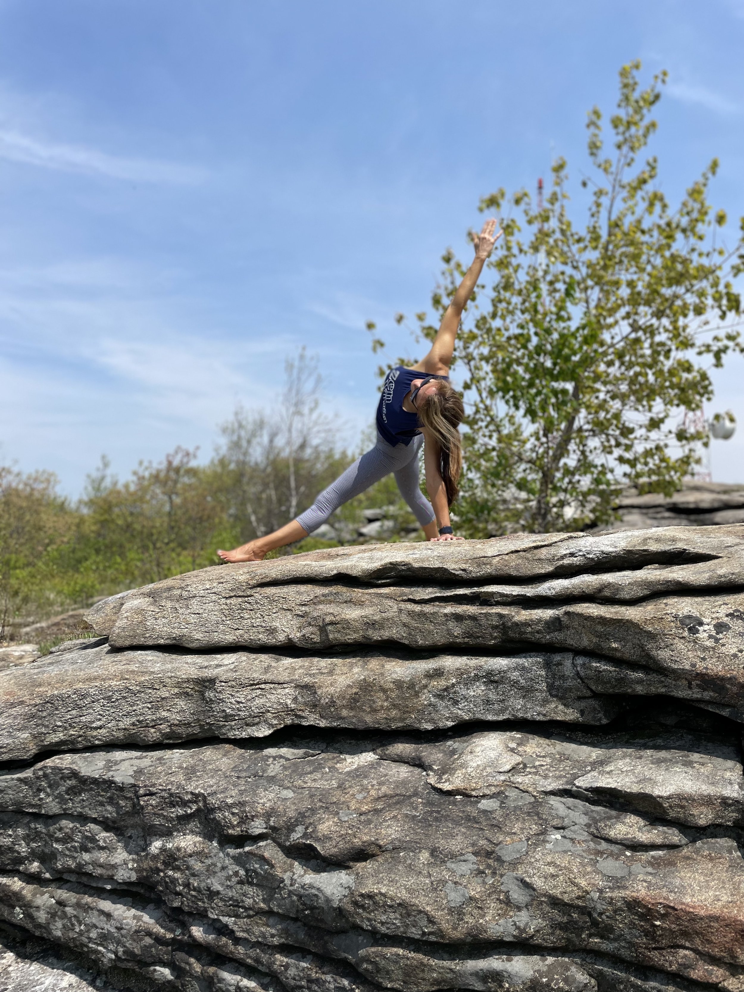 Person practicing yoga on a rocky surface with a tree and blue sky in the background.