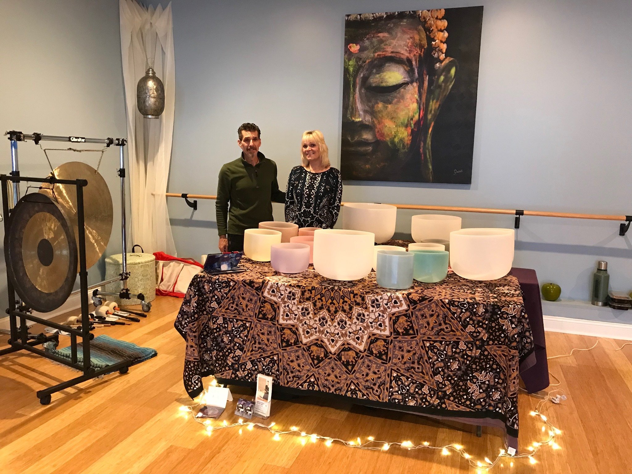 Two people standing behind a table with crystal singing bowls and a tapestry, with a gong and a large Buddha painting in the background.