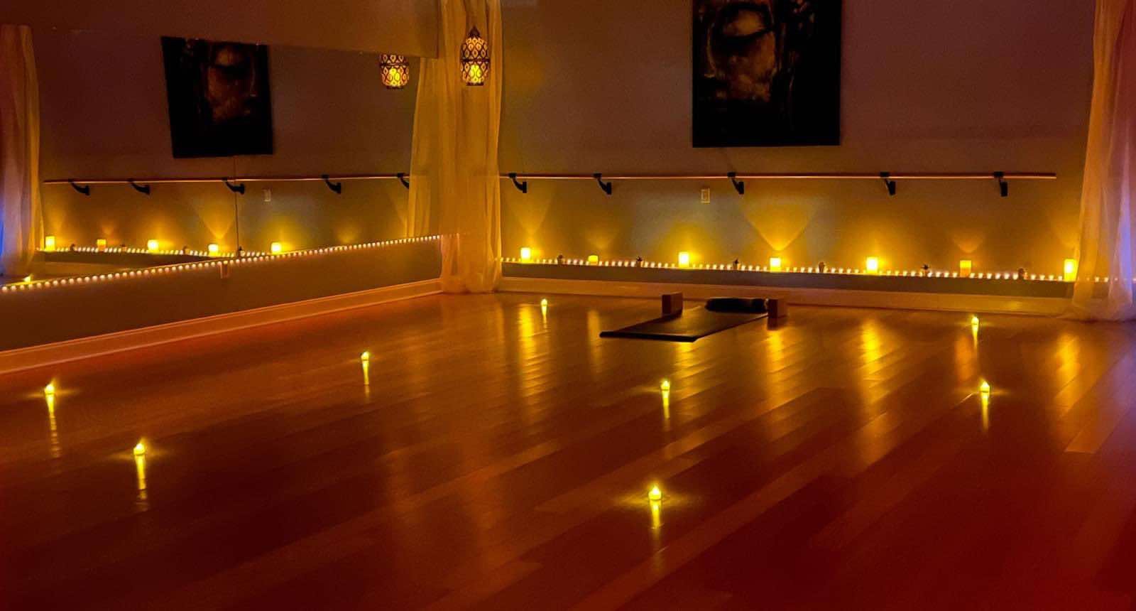 Dimly lit yoga studio with candles and a yoga mat on a wooden floor, surrounded by soft lighting and draped curtains.
