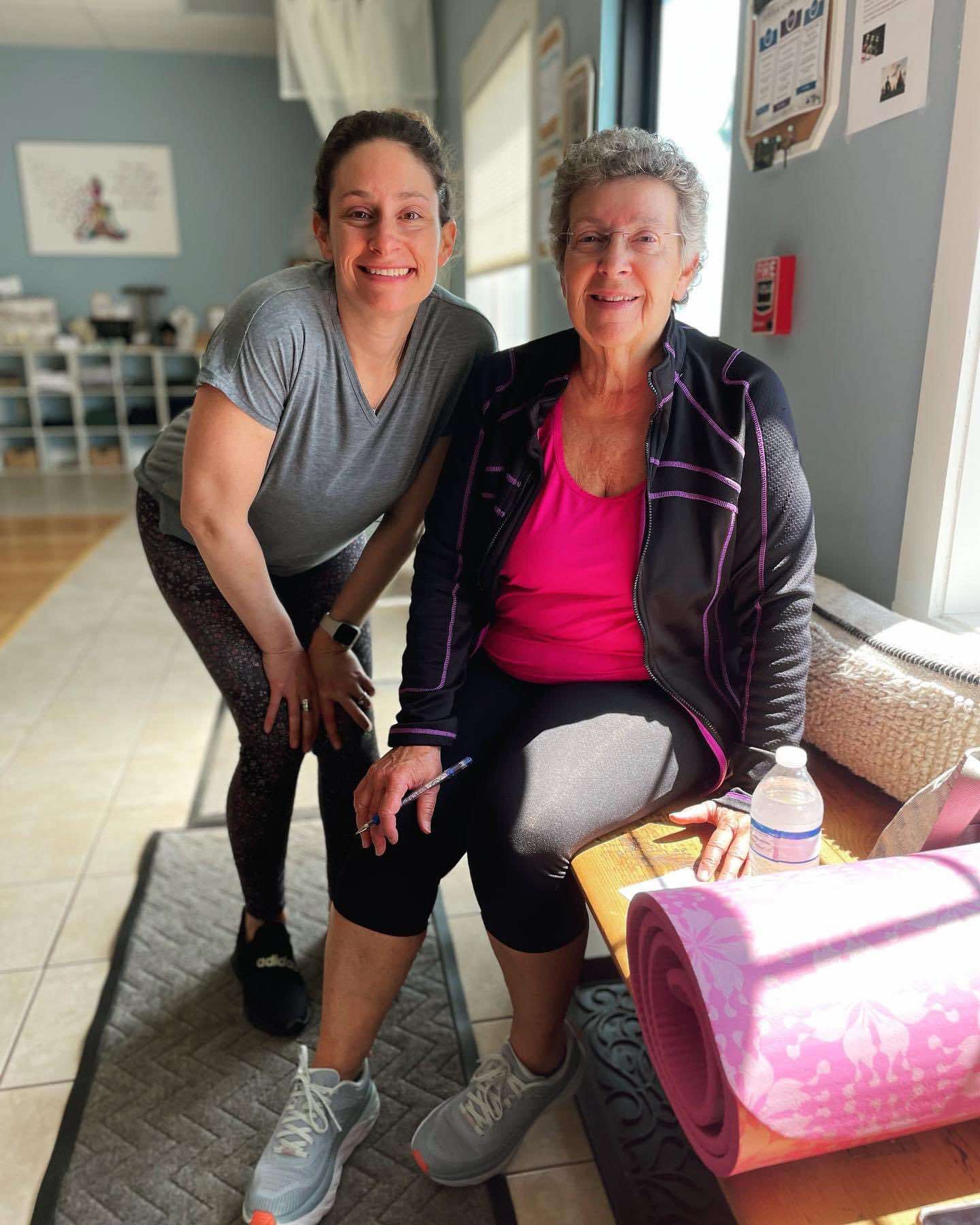 Two women in a fitness setting, one standing and one seated, both smiling. The seated woman holds a pen and has a pink yoga mat next to her. Sunlight is streaming in from a window.