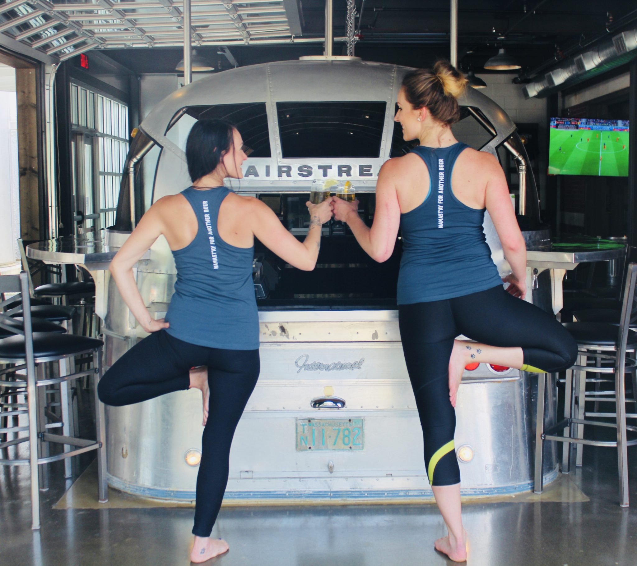 Two women wearing exercise outfits standing on one leg in tree pose, each holding a beer, in front of an Airstream trailer converted into a bar.