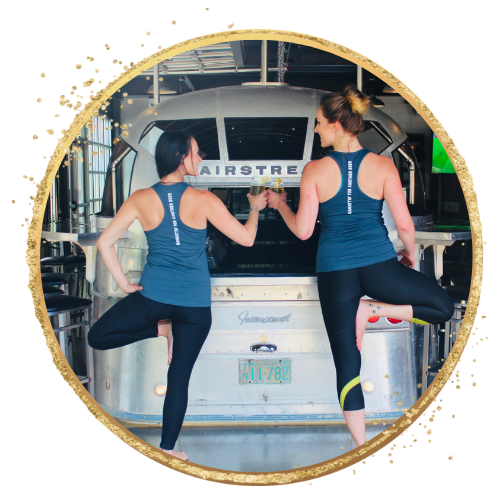 Two women in yoga poses in front of an Airstream trailer, wearing matching tank tops and leggings. They are clinking glasses in a celebratory gesture. The scene has a gold circular border with decorative sparkles.