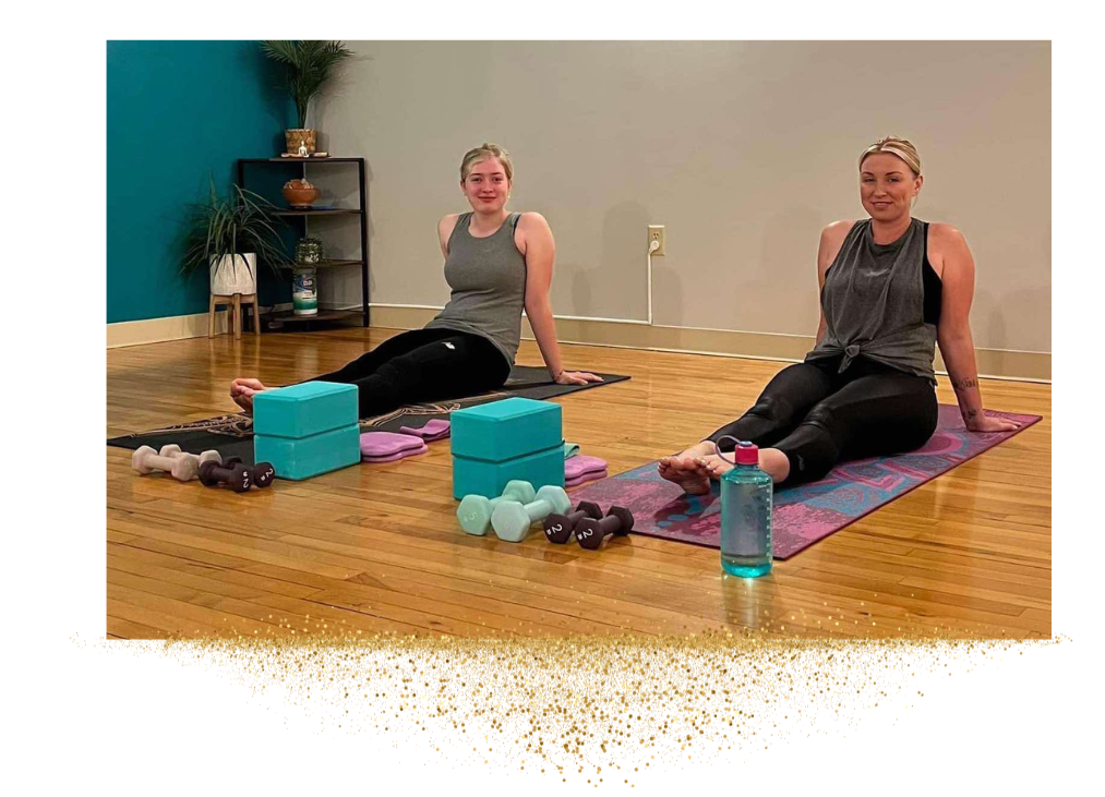 Two women sitting on yoga mats, surrounded by yoga blocks, dumbbells, and a water bottle in a fitness studio.